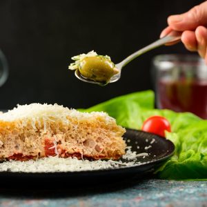 A Person Holding Stainless Steel Spoon with Green Olive Over a Plate of Bread